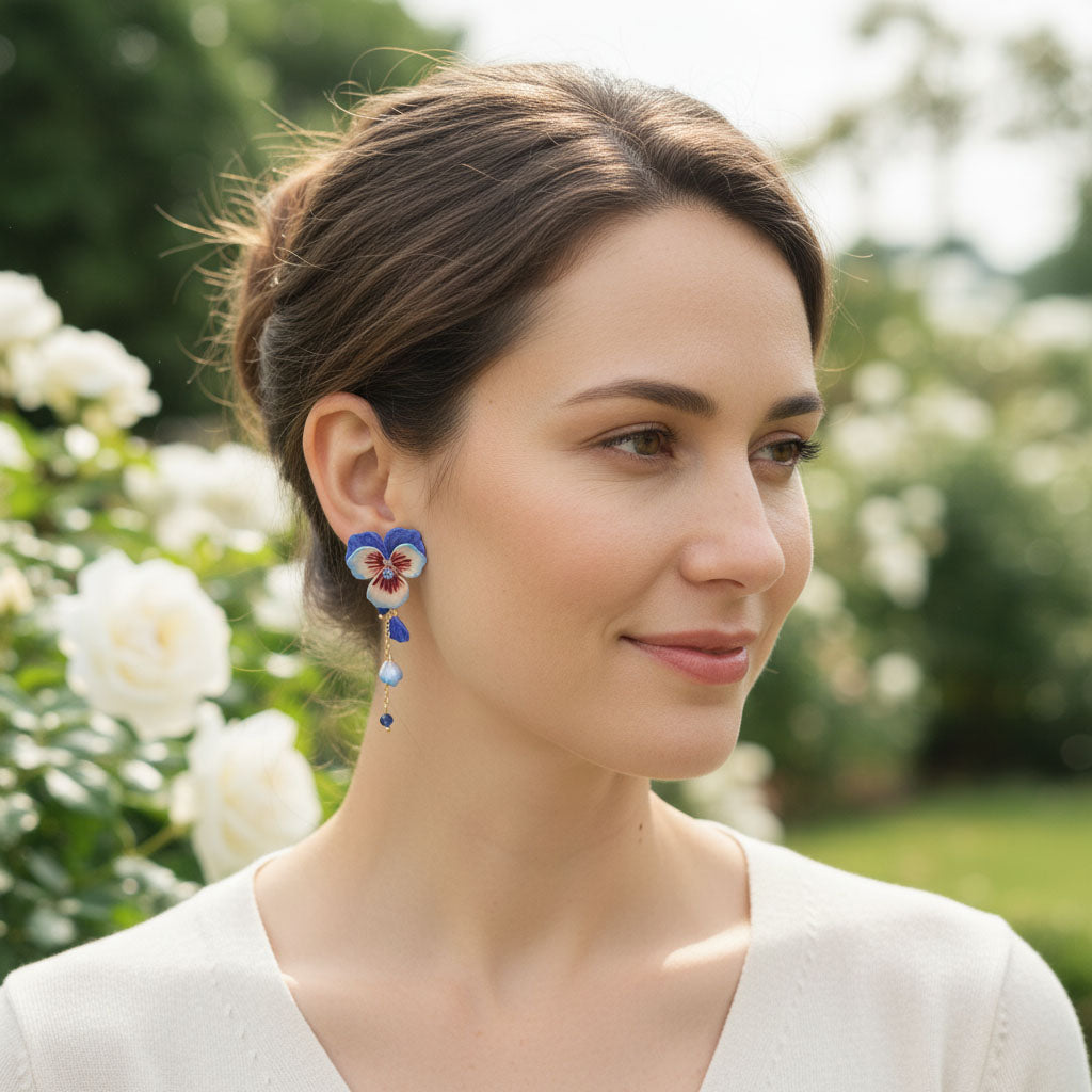 Woman wearing hand-painted blue and red pansy flower drop earrings with pearl and crystal details in a garden.