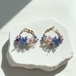 Blue and lilac flower hoop earrings with pearls displayed on stone base.