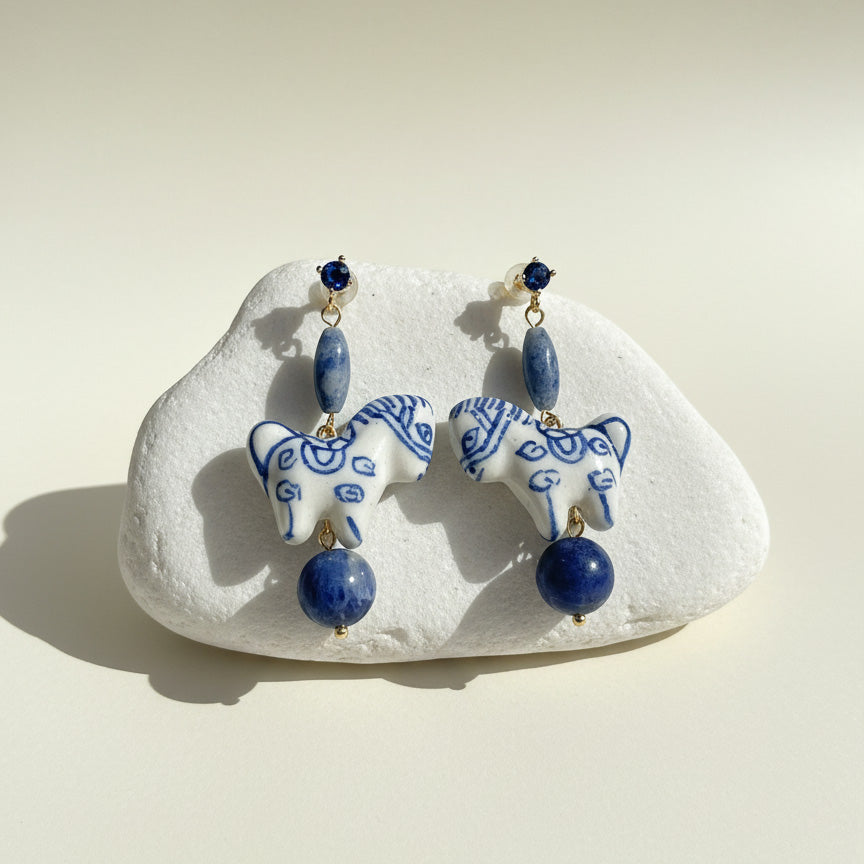 Pair of blue and white ceramic horse earrings on a white stone with a beige background