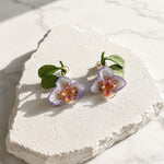 Purple orchid enamel stud earrings displayed on a textured white stone surface, featuring hand-painted petals, green resin leaves and natural stone chip accents.