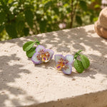 Hand-painted purple orchid enamel stud earrings styled on a natural stone surface in a sunlit garden, featuring green resin leaves and semi-precious stone chip accents.