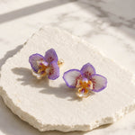 Handpainted purple orchid flower stud earrings on textured white stone surface