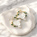 White porcelain flower hoop earrings with hand-painted green enamel leaves on a marble surface
