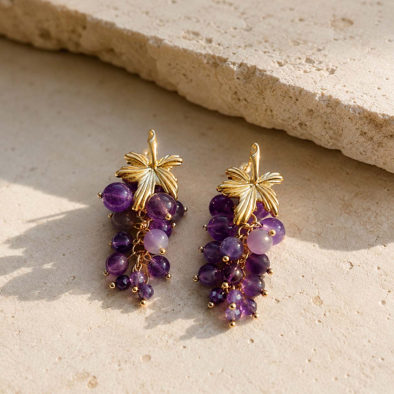 Natural amethyst grape cluster earrings with 18K gold-plated brass resting on a textured limestone surface.