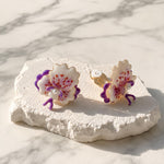 Purple orchid clip-on earrings with hand-painted enamel petals displayed on a textured white stone surface.