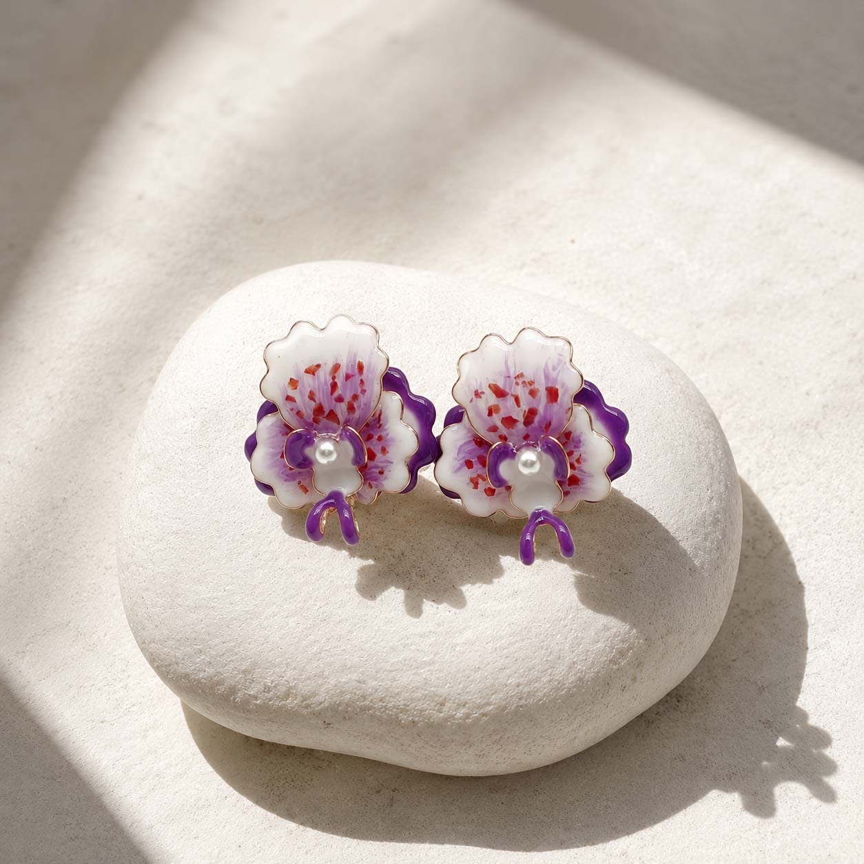 A pair of handmade orchid stud earrings with white petals, purple accents, and red speckles, resting on a smooth white stone under warm sunlight.