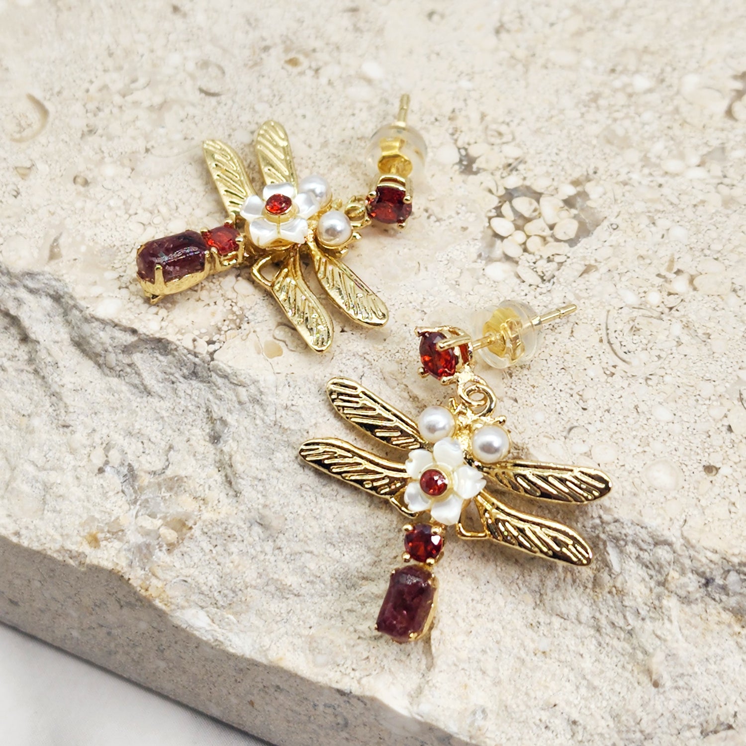 Gold dragonfly earrings with red stones, pearls, and mother-of-pearl flower accents displayed on a light stone surface.