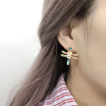 A model wearing the gold dragonfly earrings with green stones and mother-of-pearl flower accents, captured in close-up to show the earring in use.