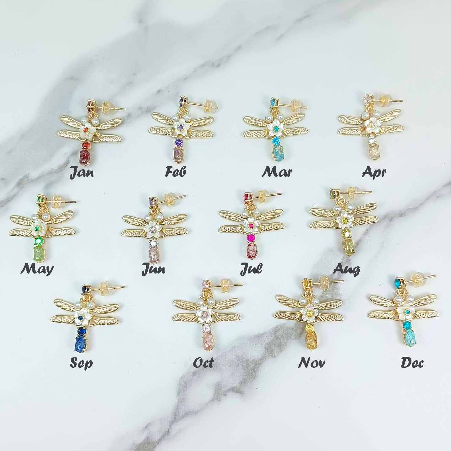 Multiple variations of the gold dragonfly earrings, each with different birthstone colors, laid out on a marble-like background.