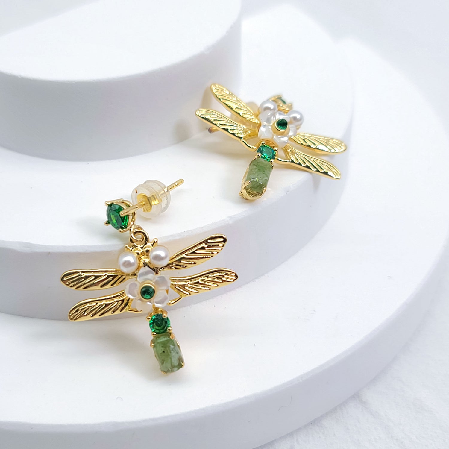 Gold dragonfly earrings with green semi-precious stones, pearls, and mother-of-pearl flowers on a white display background.
