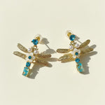 Dragonfly-shaped earrings with gold, blue, and pearl details on a beige background