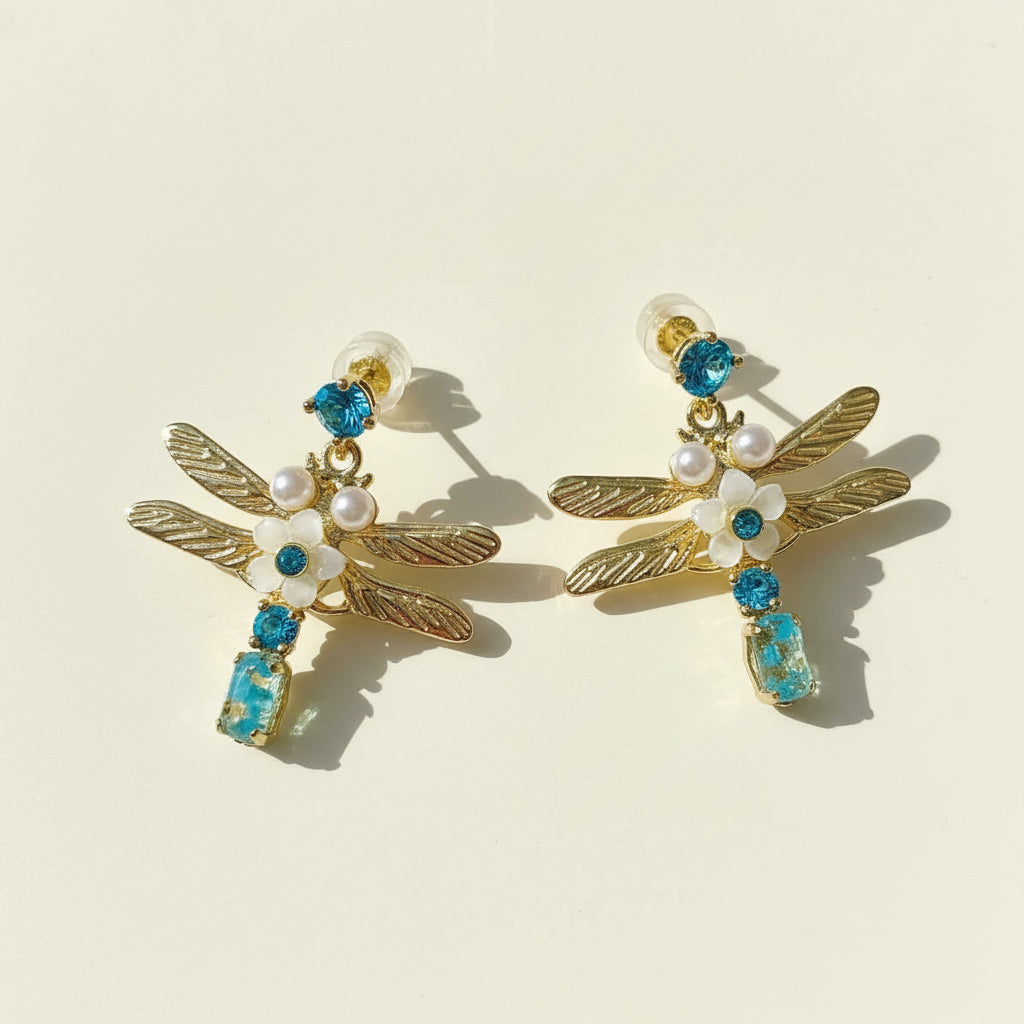 Dragonfly-shaped earrings with gold, blue, and pearl details on a beige background