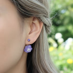Close-up of an ear wearing purple floral earrings with a blurred natural background