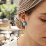 Close-up of a person wearing blue floral earrings with a blurred outdoor background
