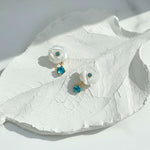 Pair of earrings with white floral designs and blue gemstones on a white leaf