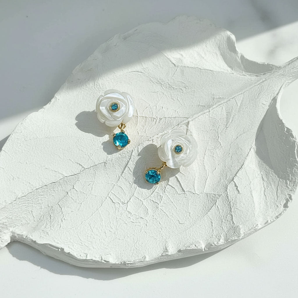 Pair of earrings with white floral designs and blue gemstones on a white leaf