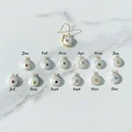 Set of pearl earrings with colorful gemstones, each labeled for a month on a marble background.