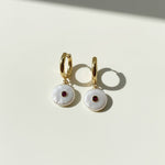 Gold hoop earrings with white circular charms and red stones on a light background