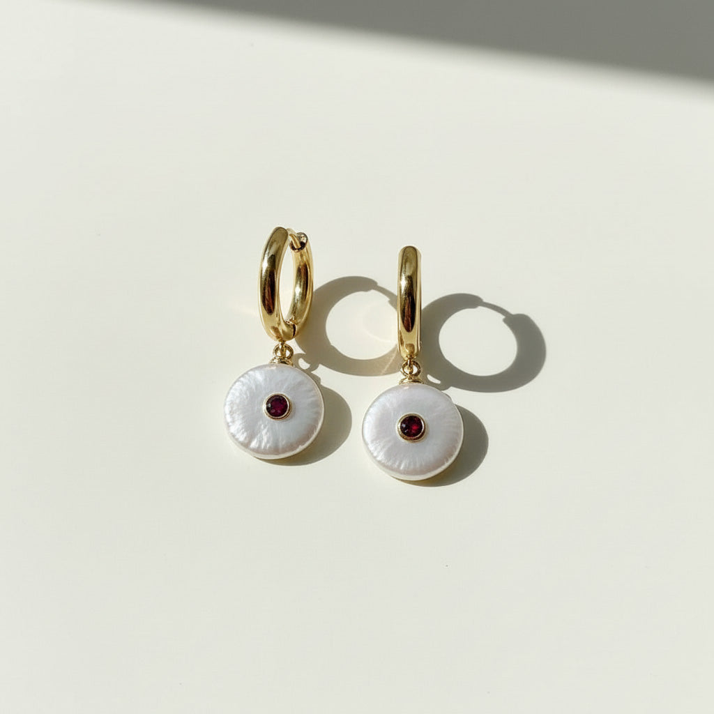 Gold hoop earrings with white circular charms and red stones on a light background