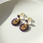 Gold and purple beaded dangle earrings with circular woven design and crystal studs on white background.