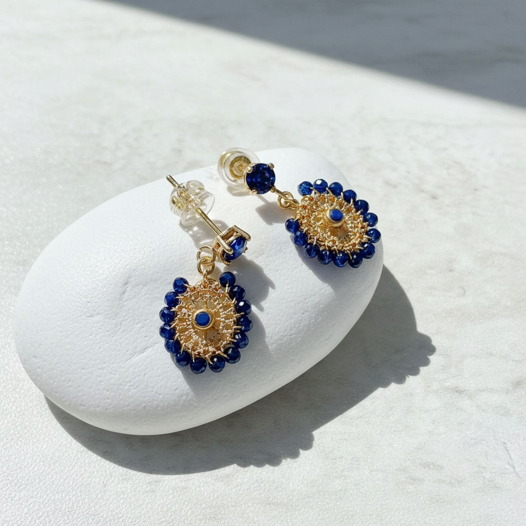 Gold and navy blue beaded drop earrings with woven circular design displayed on white stone background.