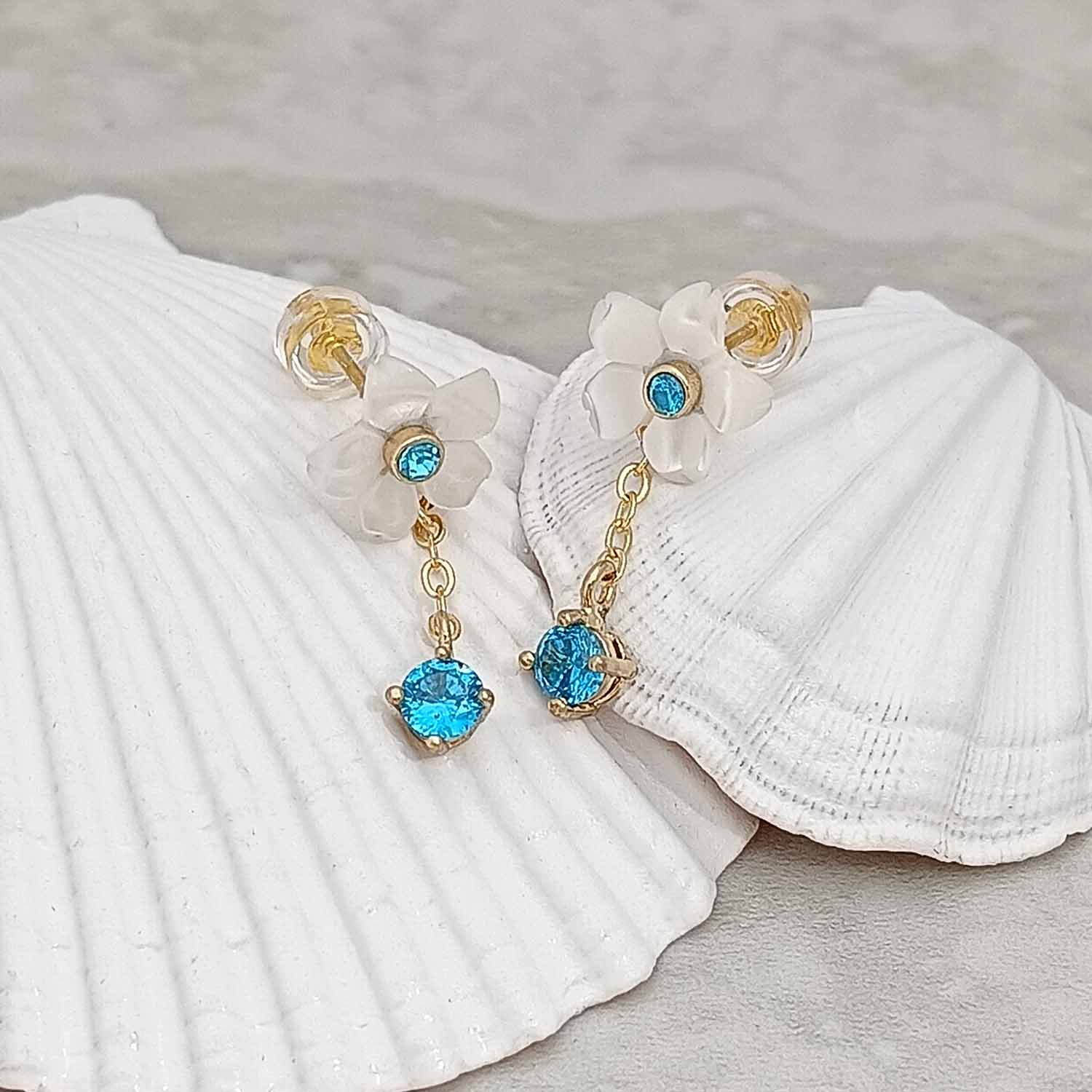 Mother of pearl floral stud earrings with blue cubic zirconia centers and matching crystal drop on gold-plated chain, styled on white seashell background