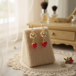 Pair of earrings with strawberry and flower designs on a beige stand.