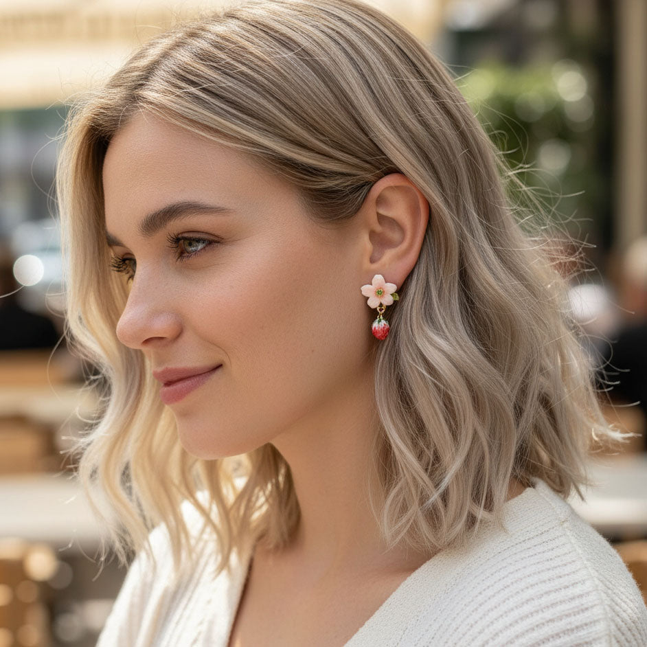 “Model wearing a handcrafted strawberry-themed earring featuring a pink flower stud and dangling red strawberry charm, photographed outdoors in natural light