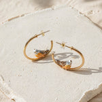 Gold hoop earrings with bird designs on a textured beige surface