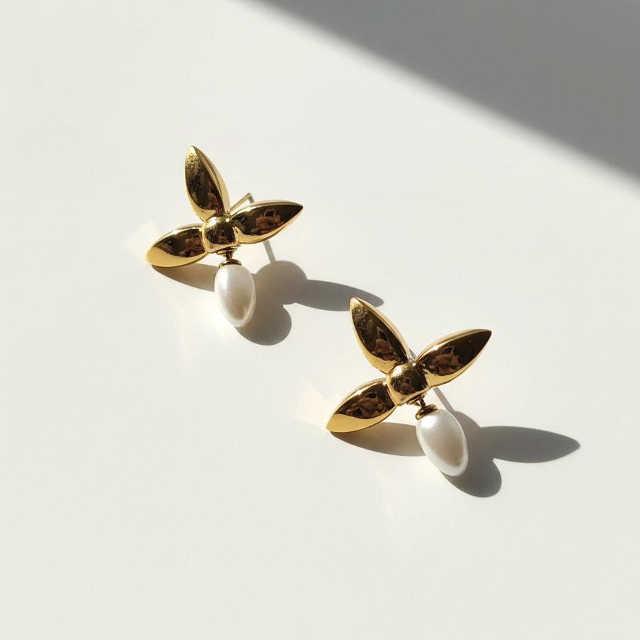 Gold floral stud earrings with three-petal design and teardrop pearl dangle, photographed in natural sunlight