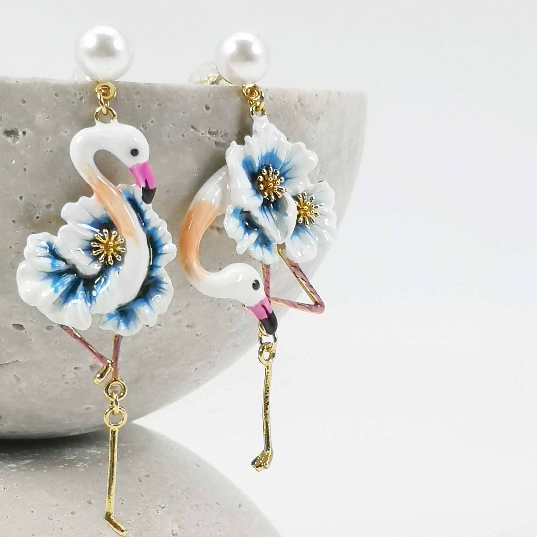 Blue floral flamingo enamel drop earrings with pearl studs, displayed on curved stone background.