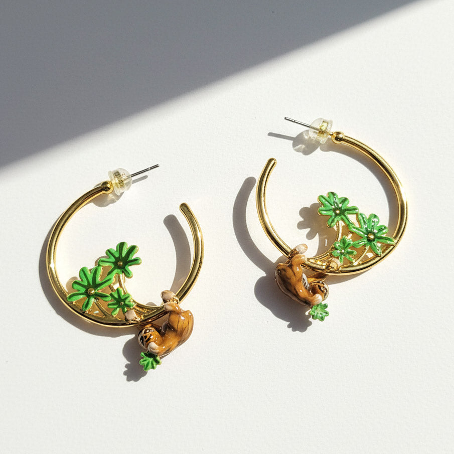 Gold hoop earrings with green leaf and brown sloth charms on a white background