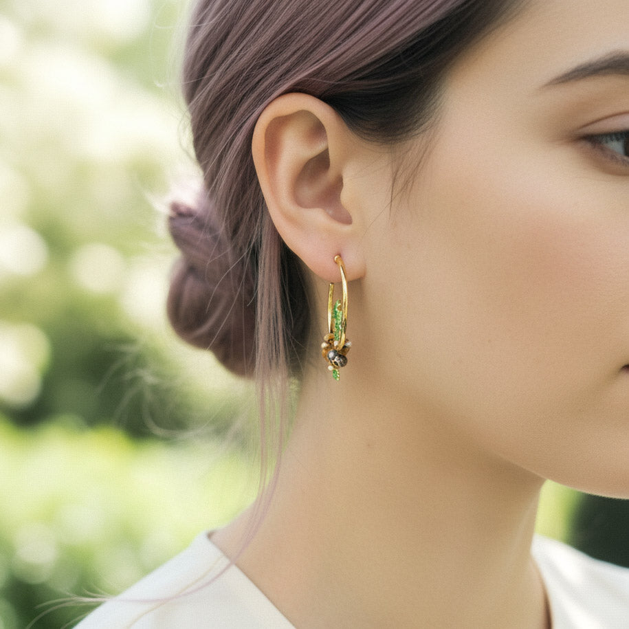Close-up of a woman wearing gold hoop earrings with green gemstones against a blurred natural background.