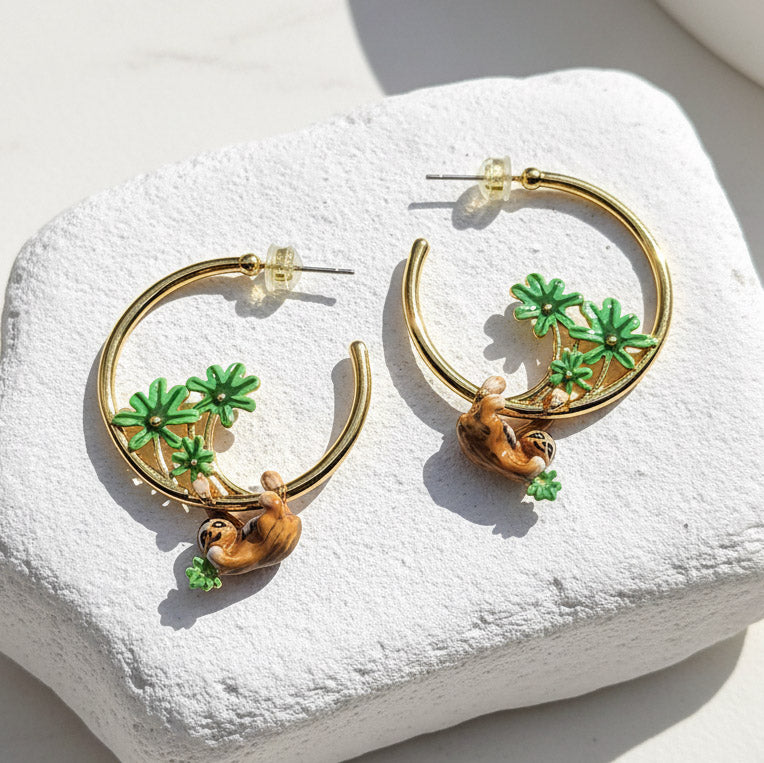 Gold hoop earrings with sloth and tree  designs on a white stone surface
