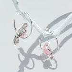 Handcrafted silver hoop earrings with pink and grey bird design, enamel painted cockatoo earrings on white branch display.