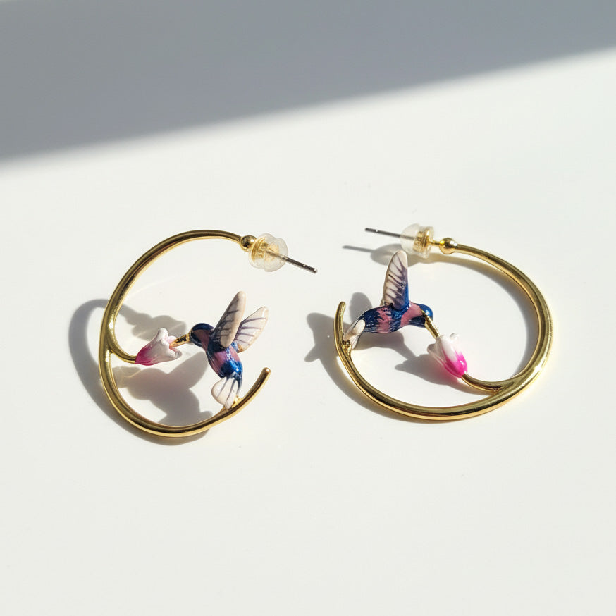 Gold hoop earrings featuring hand-painted hummingbirds sipping from pink flowers, captured in natural sunlight.
