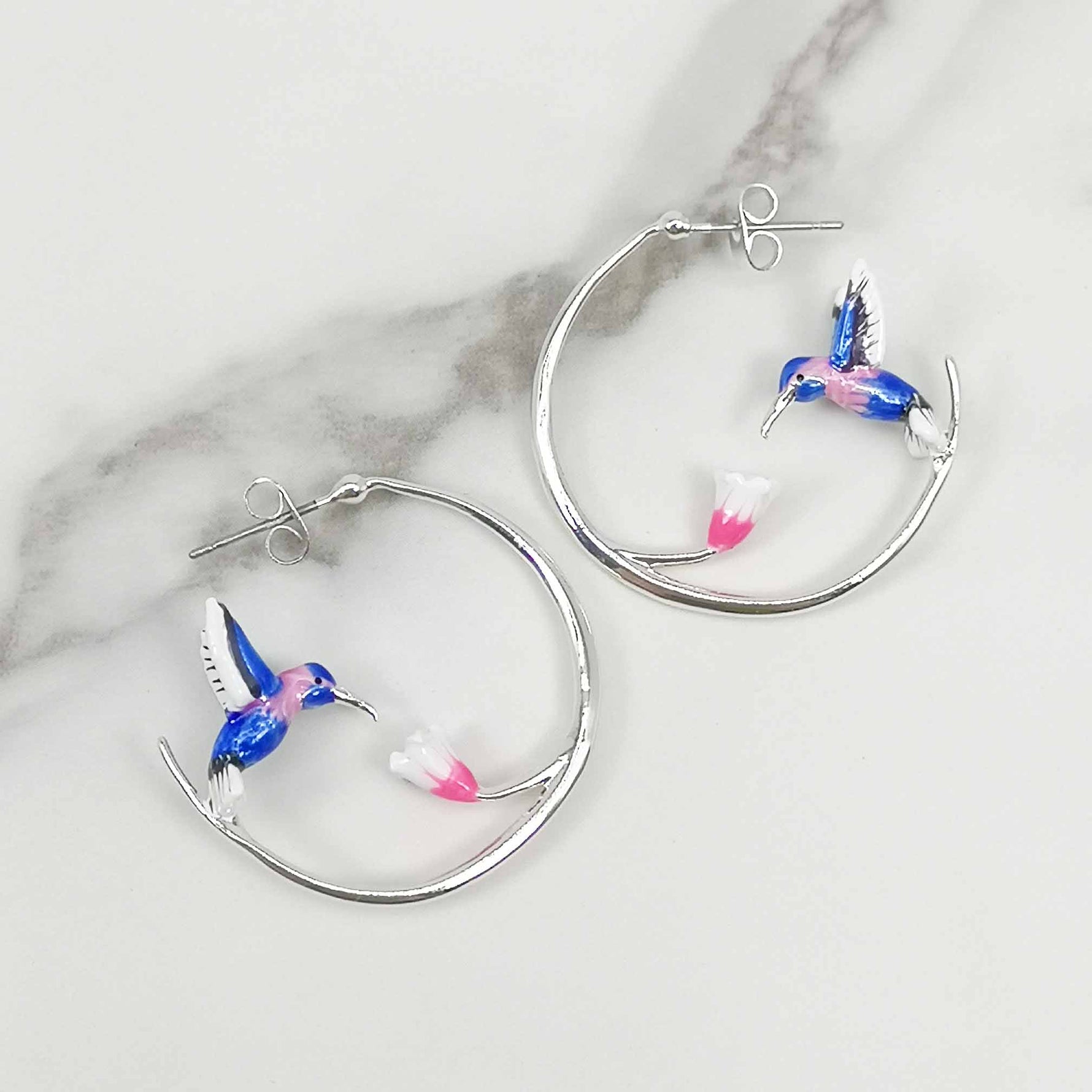 A pair of silver-plated hoop earrings featuring an enamel-painted hummingbird in shades of blue and pink. Each earring showcases a delicate hummingbird in mid-flight, feeding from a pink and white flower. The earrings have a smooth, polished finish and are photographed against a white marble-like background, emphasizing their elegant and whimsical design.