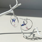 Silver hoop earrings with blue and pink enamel hummingbirds and flowers, elegant handcrafted jewelry on branch display.