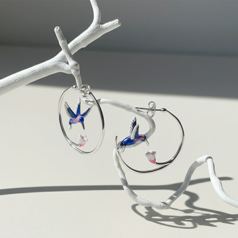 Silver hoop earrings with blue and pink enamel hummingbirds and flowers, elegant handcrafted jewelry on branch display.