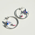 Silver hoop earrings with colorful hummingbird and pink flower design, artistic enamel jewelry on white background.