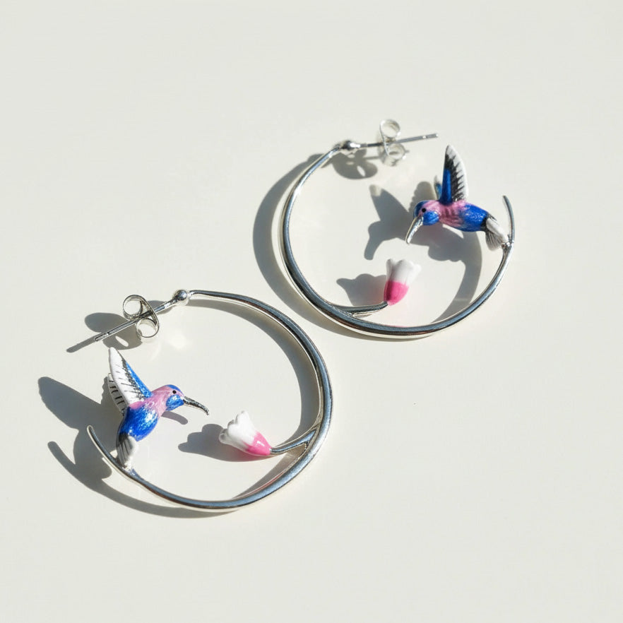 Silver hoop earrings with colorful hummingbird and pink flower design, artistic enamel jewelry on white background.