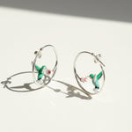 Silver hoop earrings with green enamel hummingbirds sipping from pink-white flowers, elegant handmade jewelry on light background.