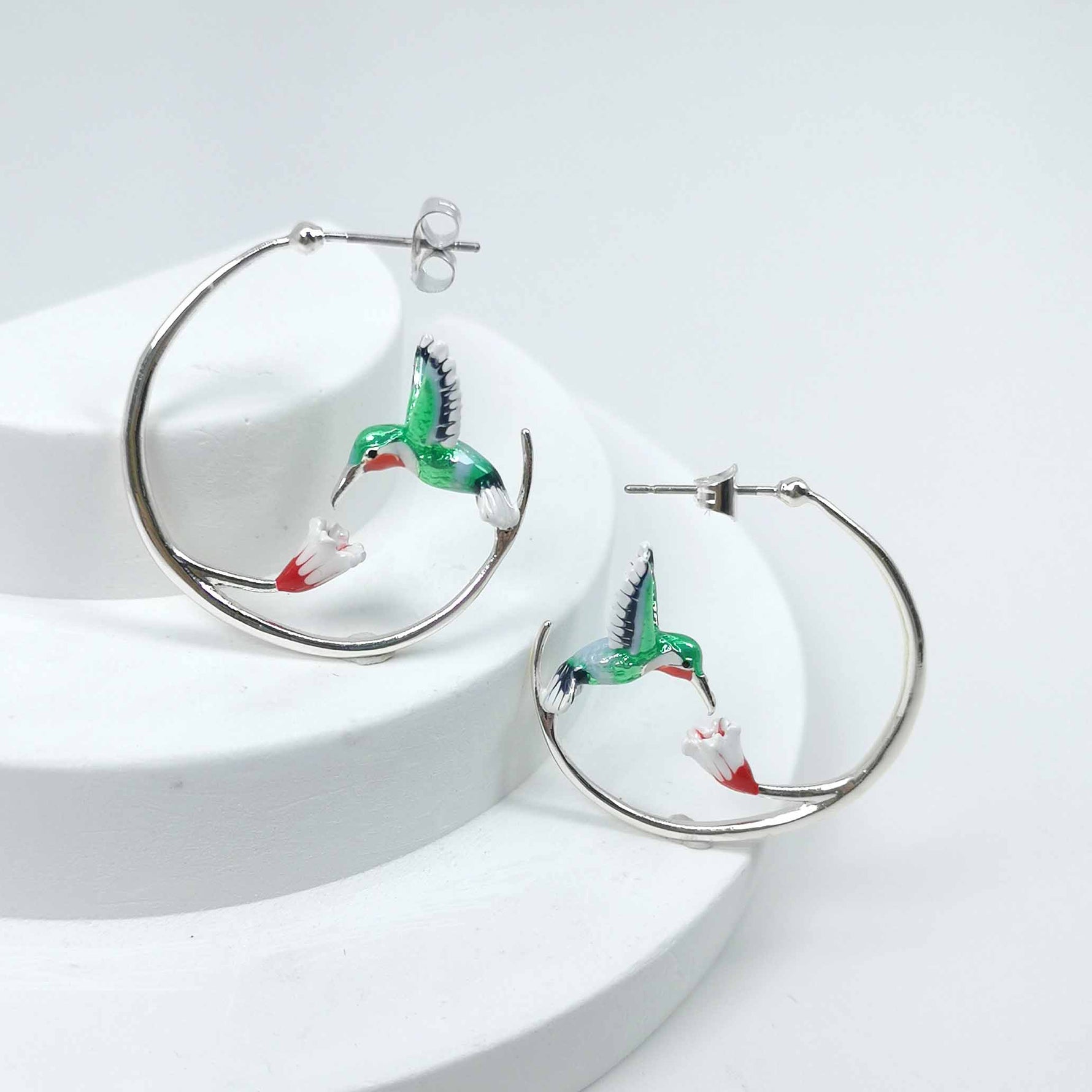 A pair of silver-plated hoop earrings featuring an enamel-painted green hummingbird in flight, feeding from a red and white flower. The earrings are displayed on a white circular stand, showcasing the vibrant colors and elegant curves of the design.