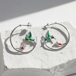 Silver hoop earrings with green enamel hummingbirds and red-white flowers, artistic nature-inspired jewelry on stone display.