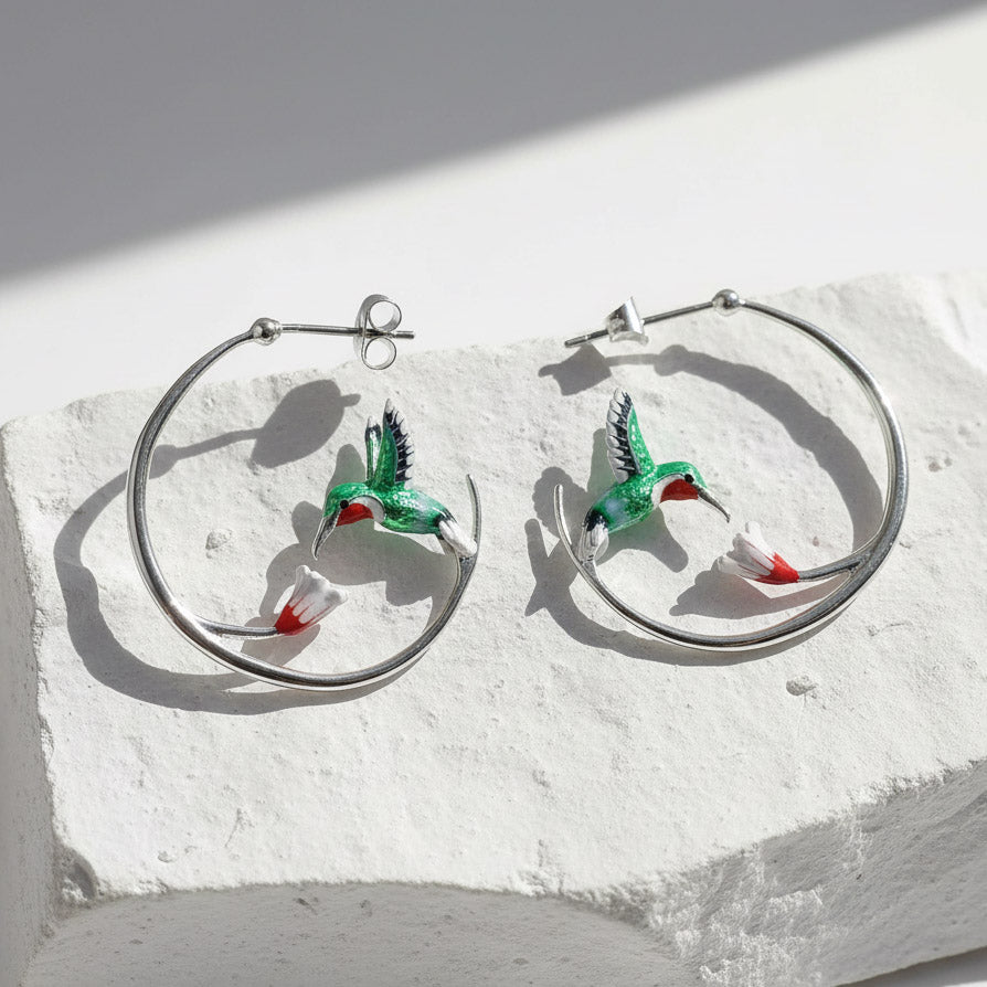 Silver hoop earrings with green enamel hummingbirds and red-white flowers, artistic nature-inspired jewelry on stone display.