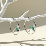 Silver hoop earrings with green enamel hummingbirds and white flowers, elegant nature-inspired jewelry on display branch
