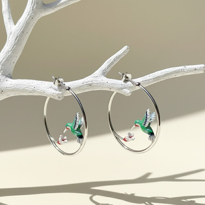 Silver hoop earrings with green enamel hummingbirds and white flowers, elegant nature-inspired jewelry on display branch