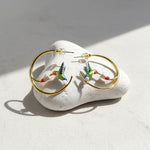 Gold hoop earrings with colorful hummingbird designs on a white stone surface.