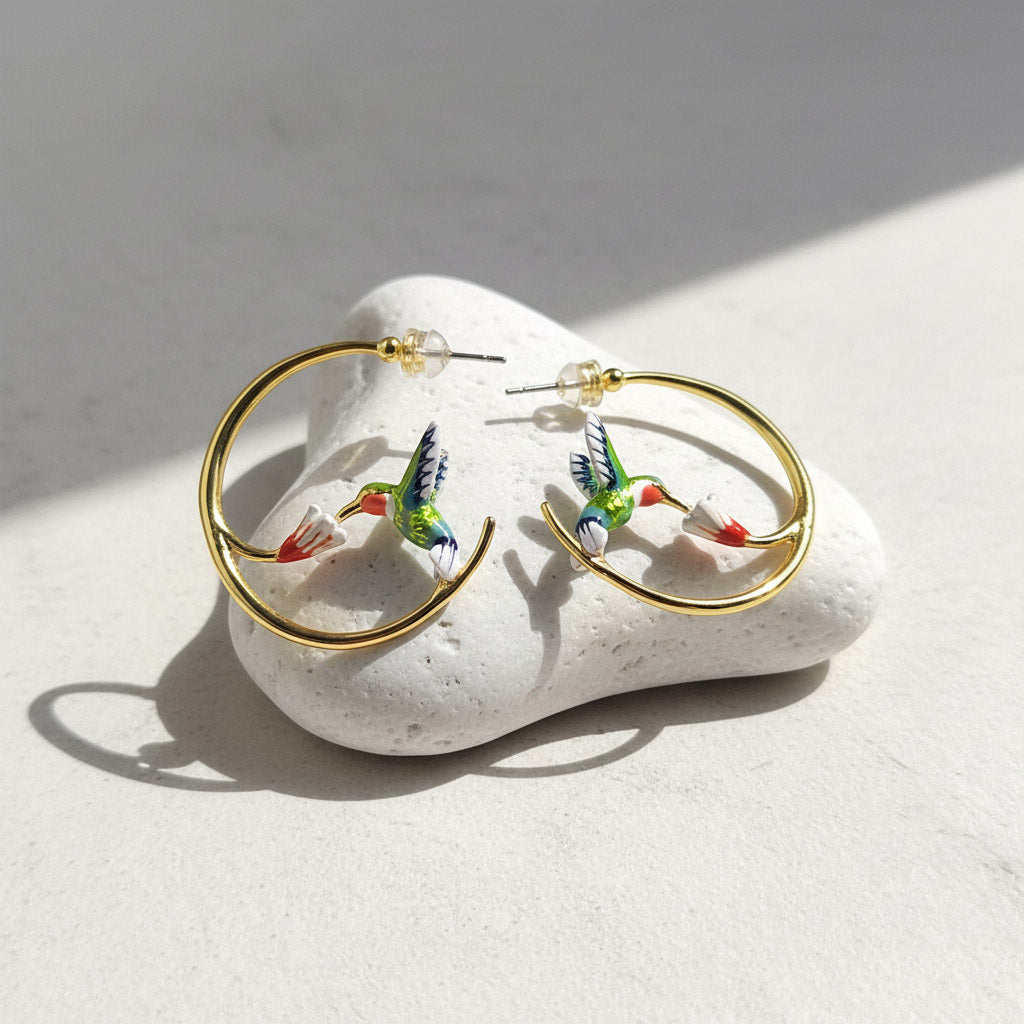 Gold hoop earrings with colorful hummingbird designs on a white stone surface.