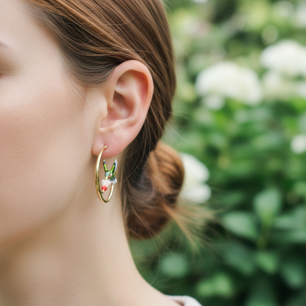 Model wearing gold hoop earrings with hand-painted hummingbird and flower design, photographed outdoors.